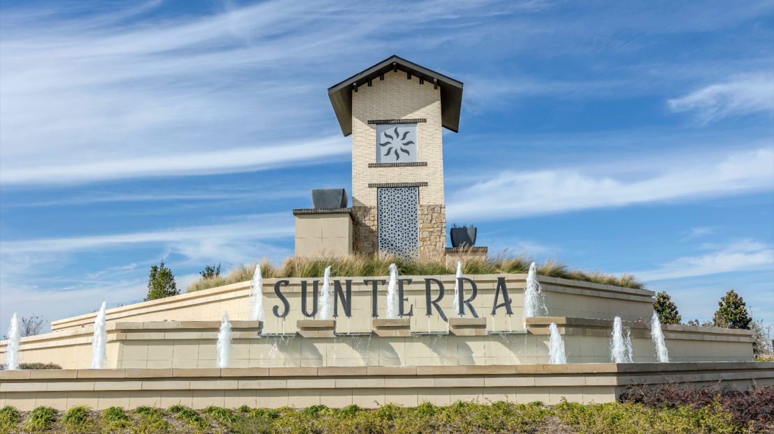 Large stone monument with a clock tower and fountains in front, displaying the word "Sunterra"—a proud feature of a top community in Texas—against a blue sky with light clouds.
