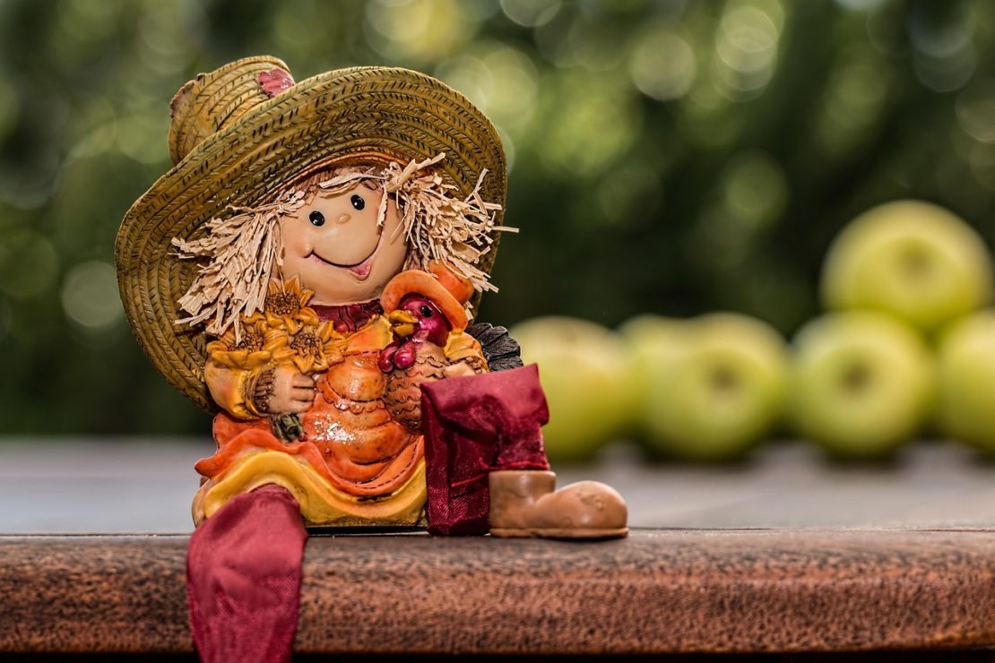 A small scarecrow figurine in autumn-themed clothing sits on a wooden surface with green apples blurred in the background.