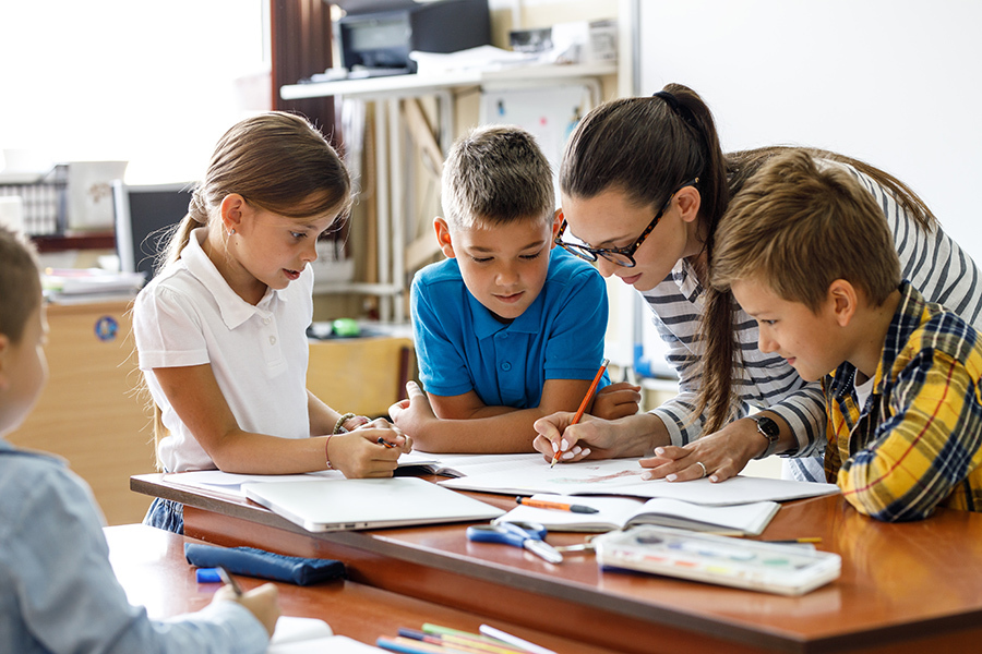 A teacher helps three elementary school children with a writing activity at a classroom table, with notebooks and art supplies visible.