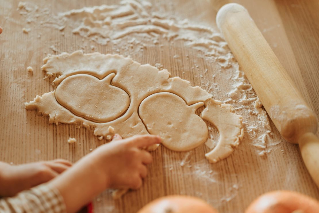 Child cutting pumpkin shapes from dough with a cookie cutter on a floured surface.