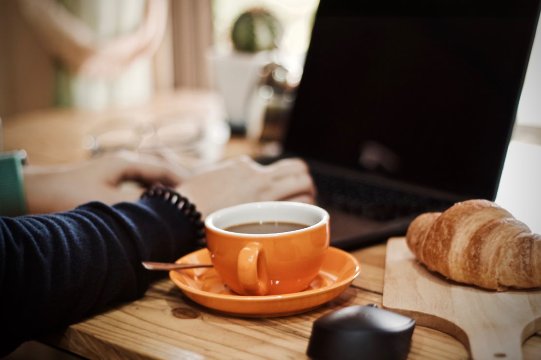 Person working on a laptop at a table with coffee in an orange cup and a croissant nearby.
