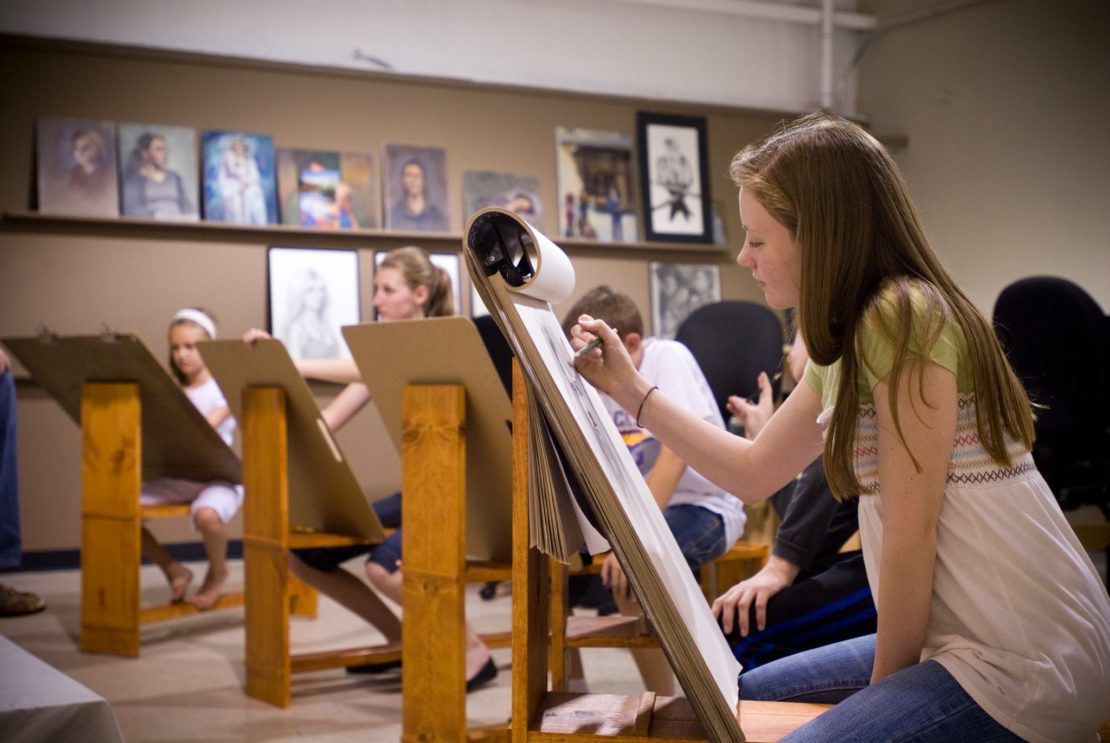 Students at summer camp sit at easels sketching in an art classroom with drawings on the wall.